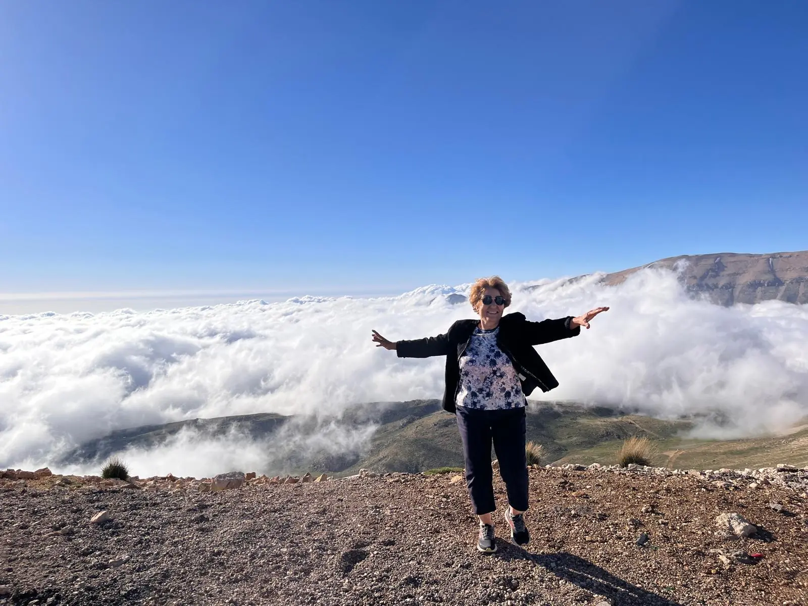 A woman smiling with arms outstretched on a mountaintop above the clouds